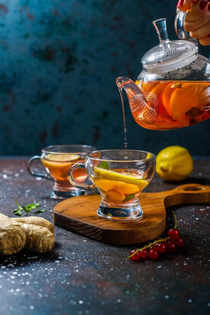 Pouring herbal tea with lemon slices into glass cup on wooden board, surrounded by fresh ginger and berries