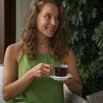 Woman in green dress smiling while holding a cup of tea, sitting on a wicker chair, surrounded by greenery