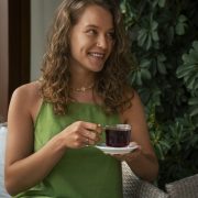 Woman in green dress smiling while holding a cup of tea, sitting on a wicker chair, surrounded by greenery